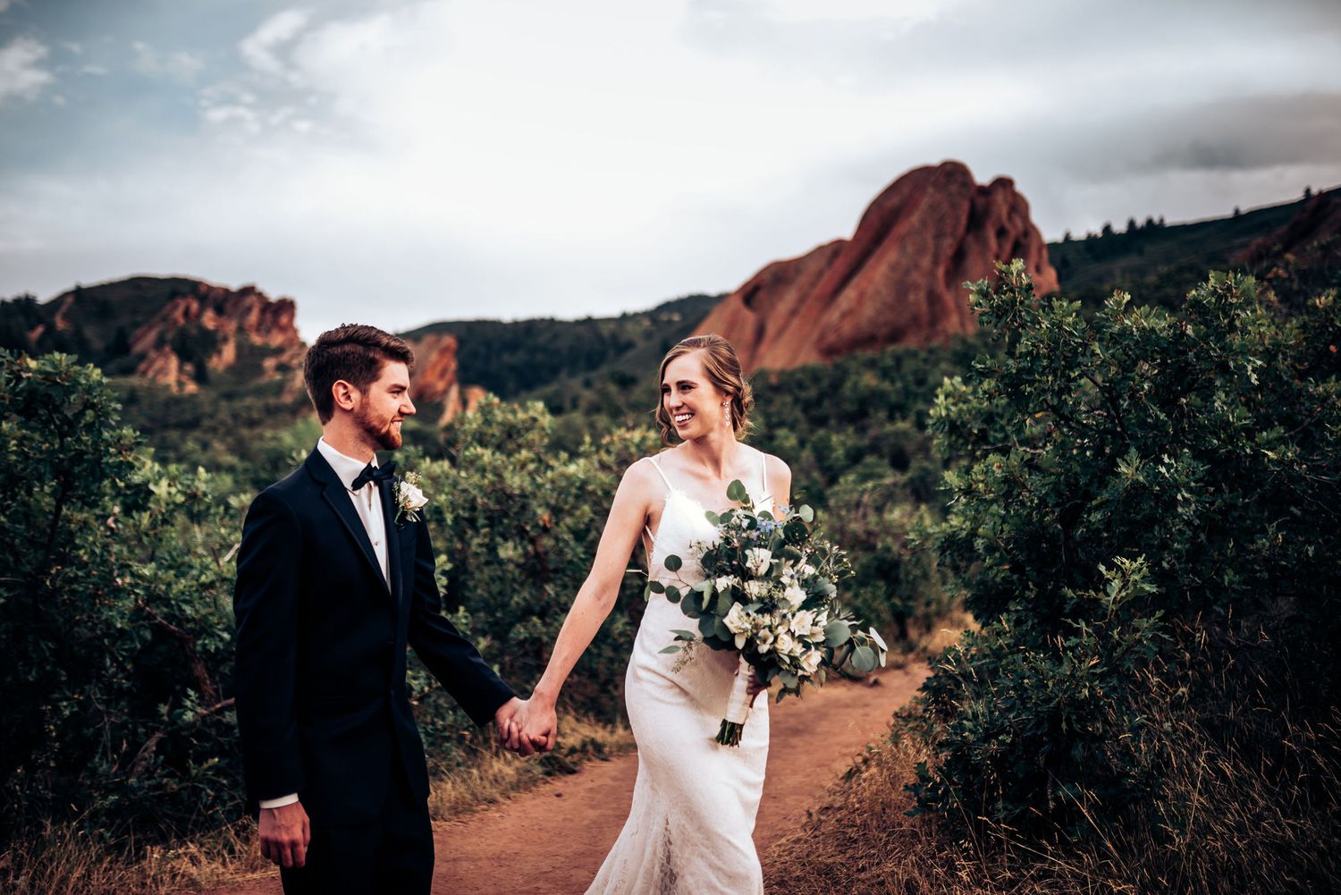 Wedding couple strolls along red rock trail with dramatic mountain formations in background.