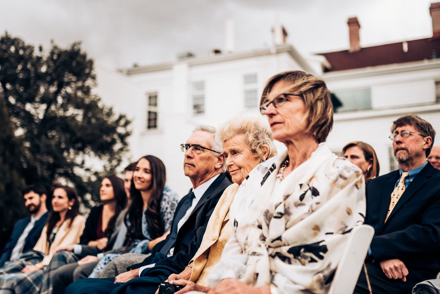 Wedding guests seated outdoors with white house visible in background.