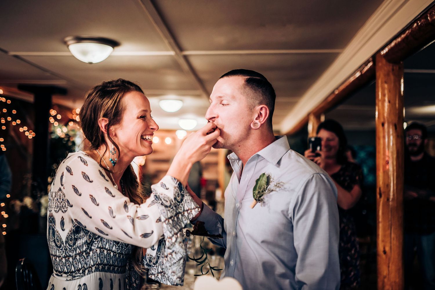 A playful cake-feeding moment during a wedding reception with guests watching.