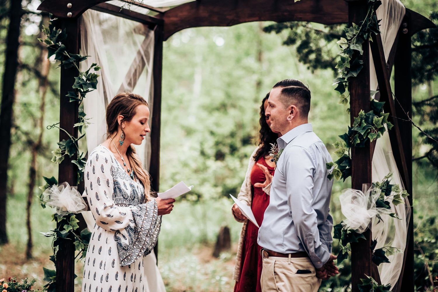 An intimate outdoor ceremony under a wooden arch decorated with greenery and white fabric.