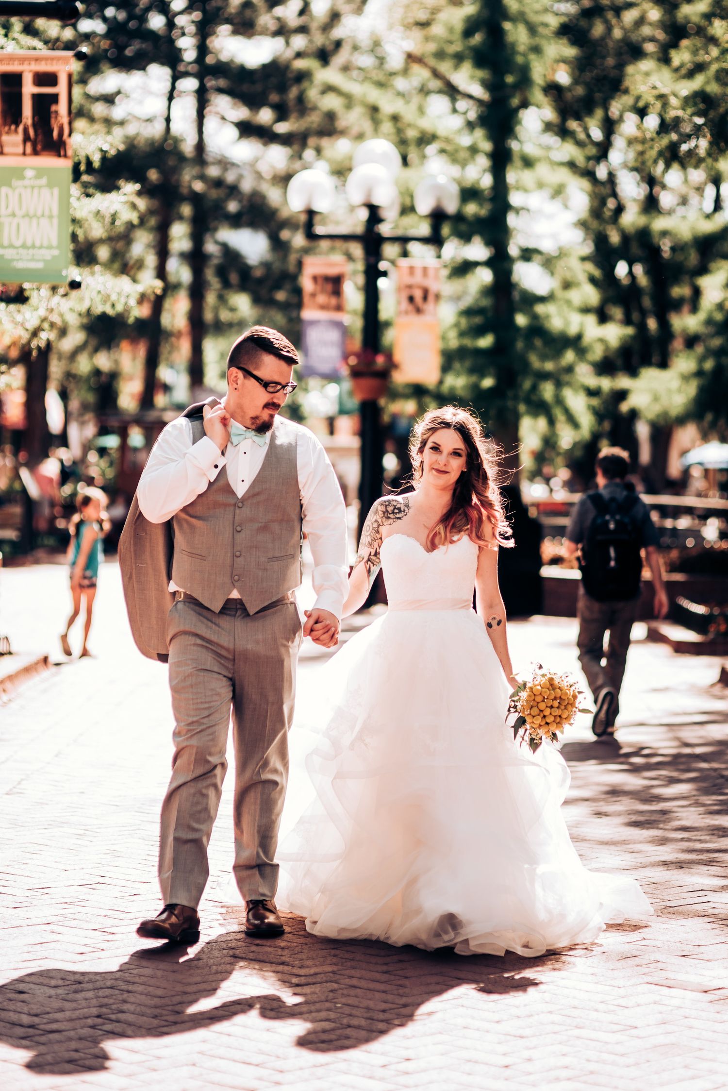 A couple walking together on a sunlit brick pathway lined with trees and banners.