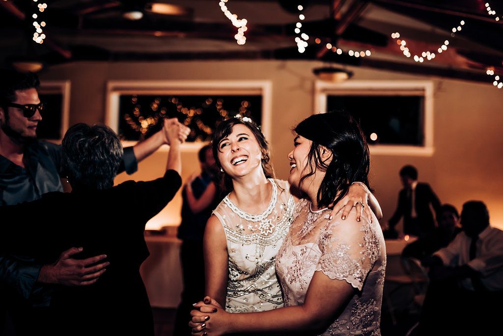 Two guests laugh joyfully while dancing at an indoor reception with twinkling lights overhead.
