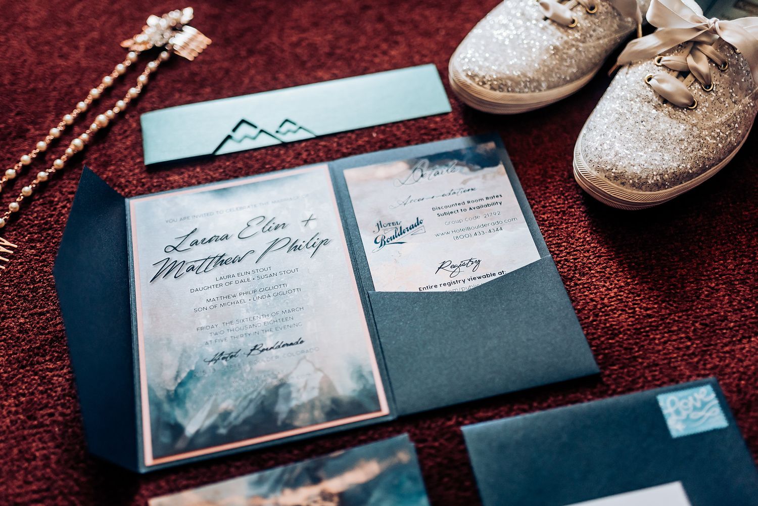 Wedding invitation suite with sparkly shoes and jewelry on burgundy surface.
