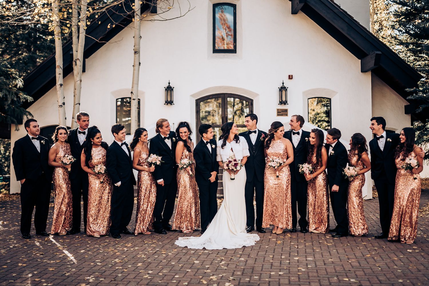Full wedding party in rose gold and black attire poses in front of white church building.