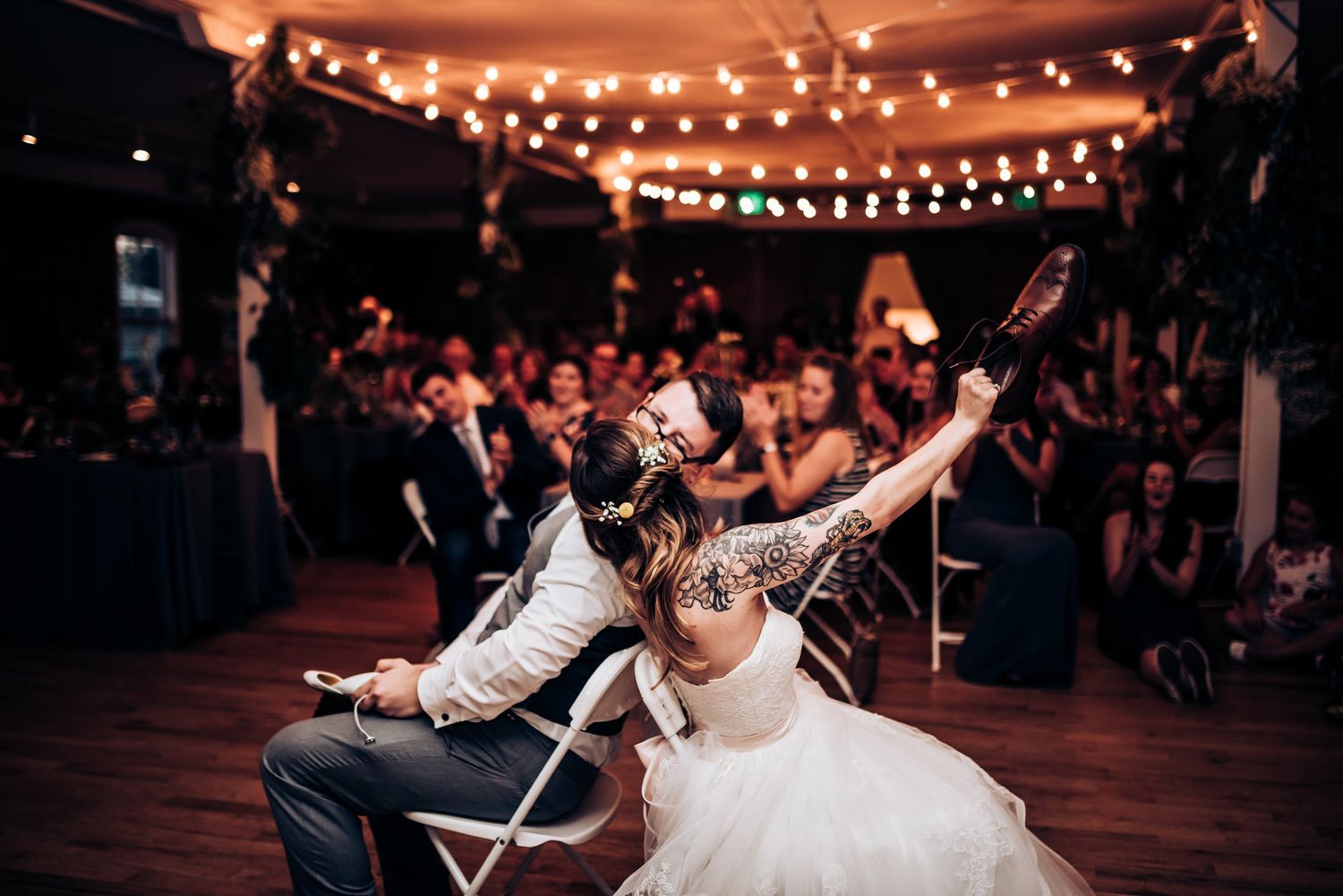 Wedding celebrants lean back in matching chairs under warm string lights at a lively reception.