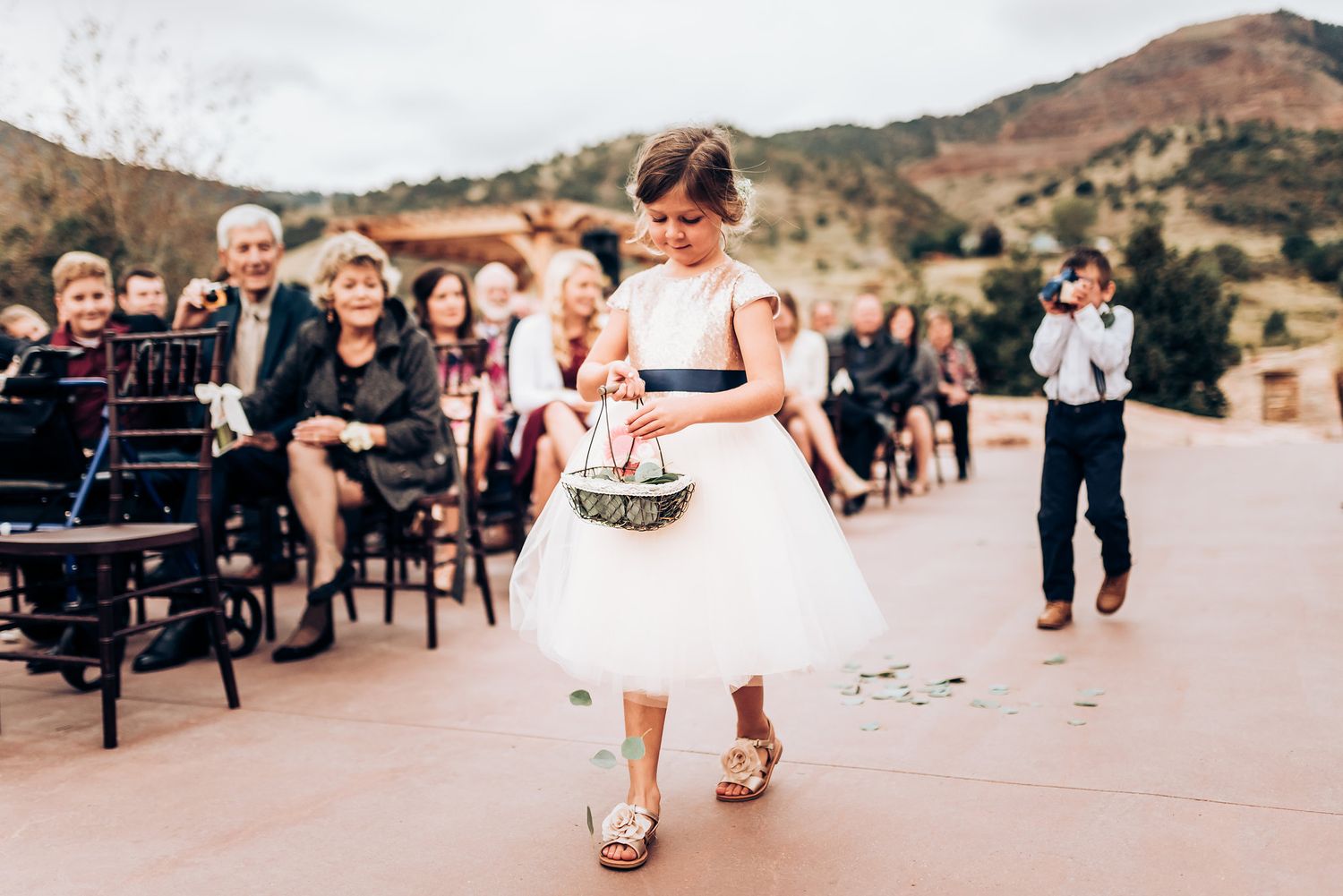 A flower girl in a white dress spreads petals during a wedding ceremony with mountains in background.