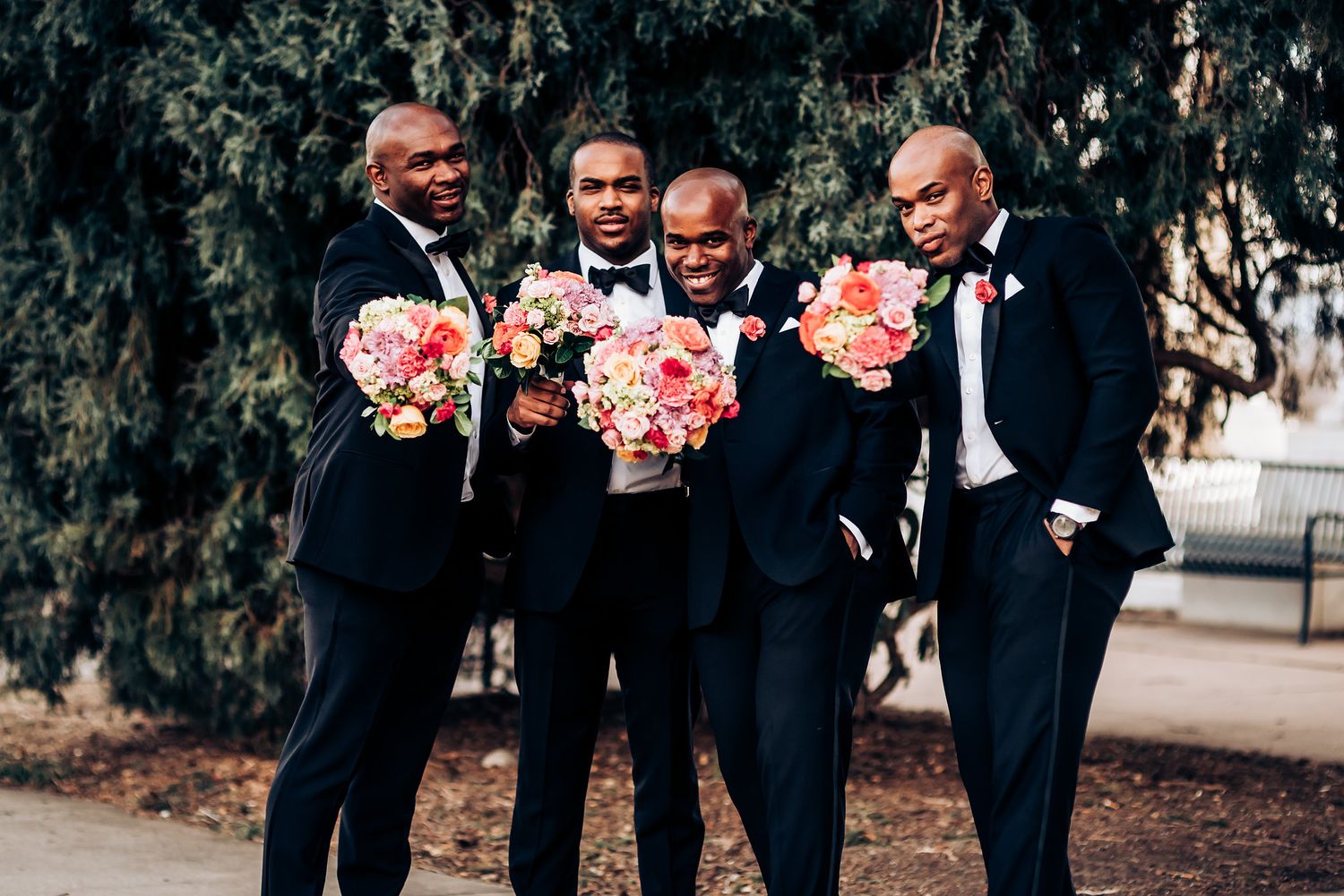 Groomsmen in navy suits holding pink and white bouquets smile together outdoors.