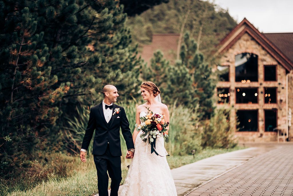 A wedding couple walks together on a mountain path with a rustic lodge in the background.