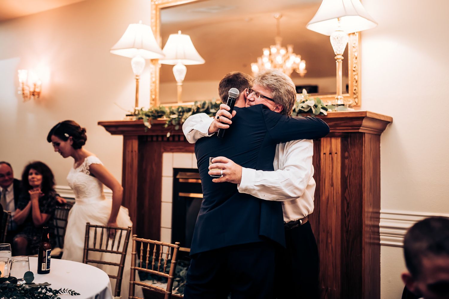 Emotional embrace shared during wedding reception toasts near an elegant fireplace.