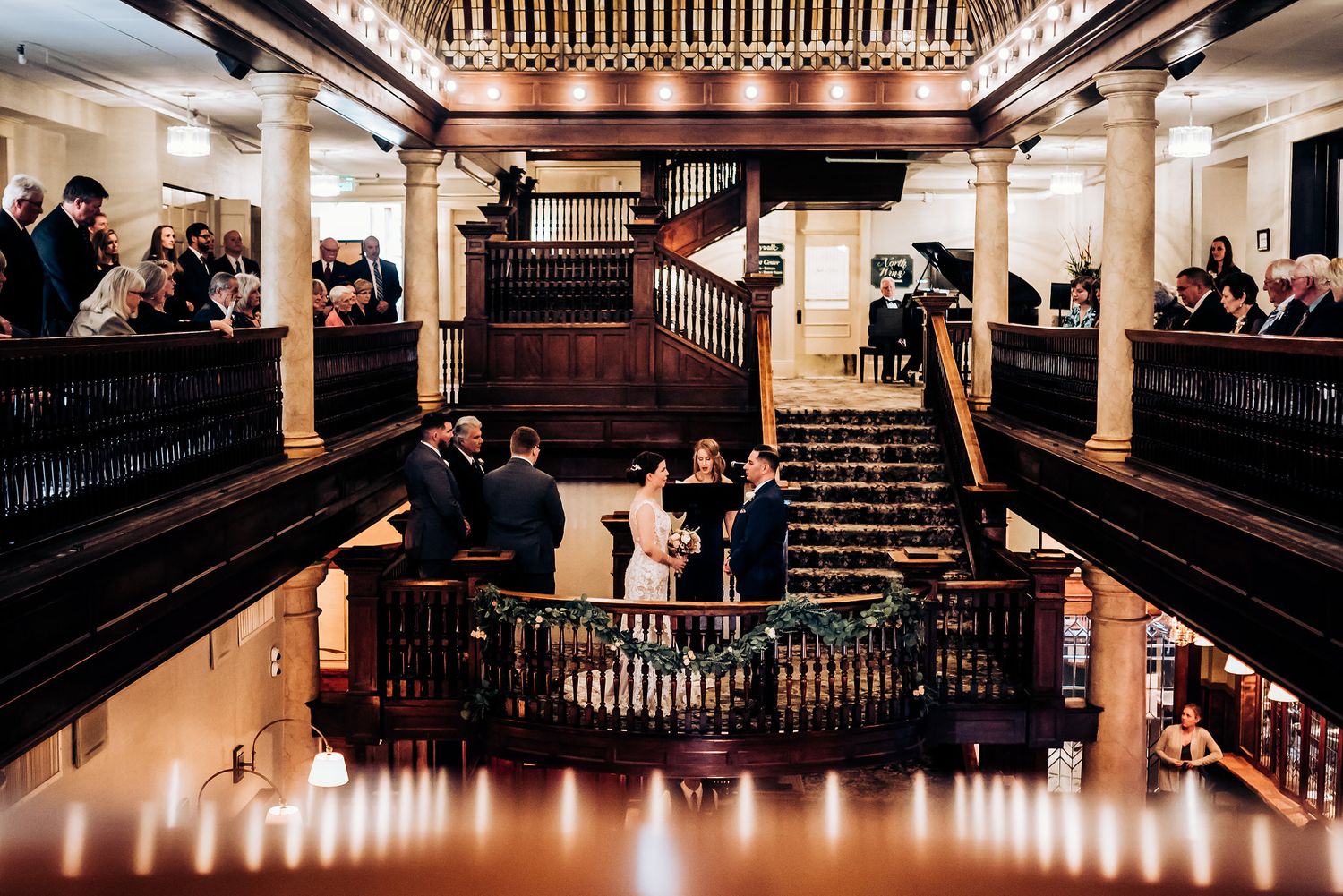 A grand wooden staircase ceremony viewed from above with guests watching from balcony levels.