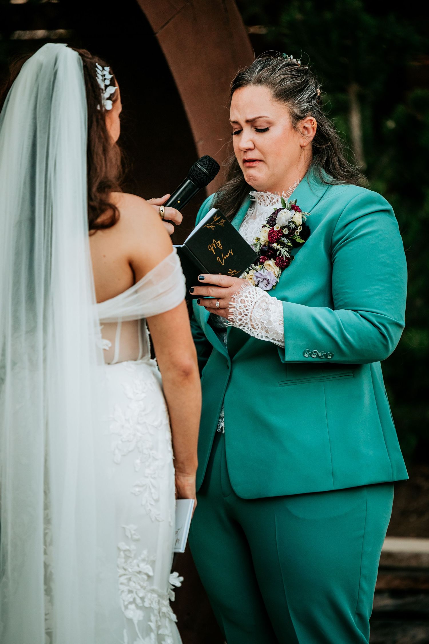 Emotional partner in teal suit reading heartfelt wedding vows from book at outdoor ceremony.