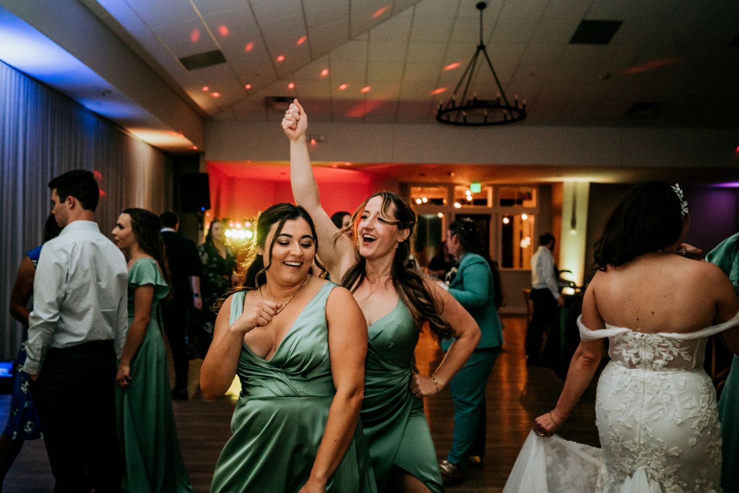 Two bridesmaids in emerald green satin dresses laugh and dance together at a wedding reception.
