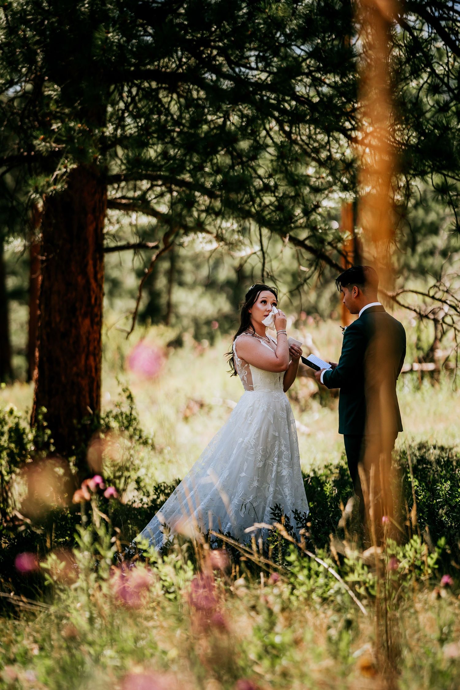 A wedding couple embraces in a sunlit forest with pink wildflowers in the foreground.