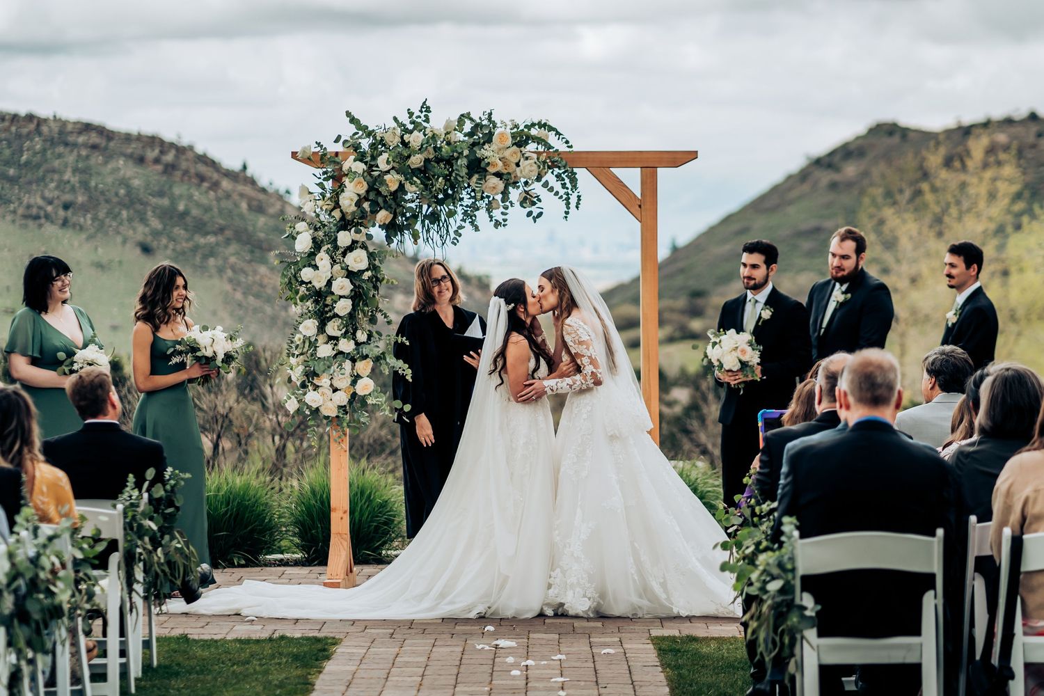 Outdoor wedding ceremony with mountain backdrop and wooden arch decorated with greenery.