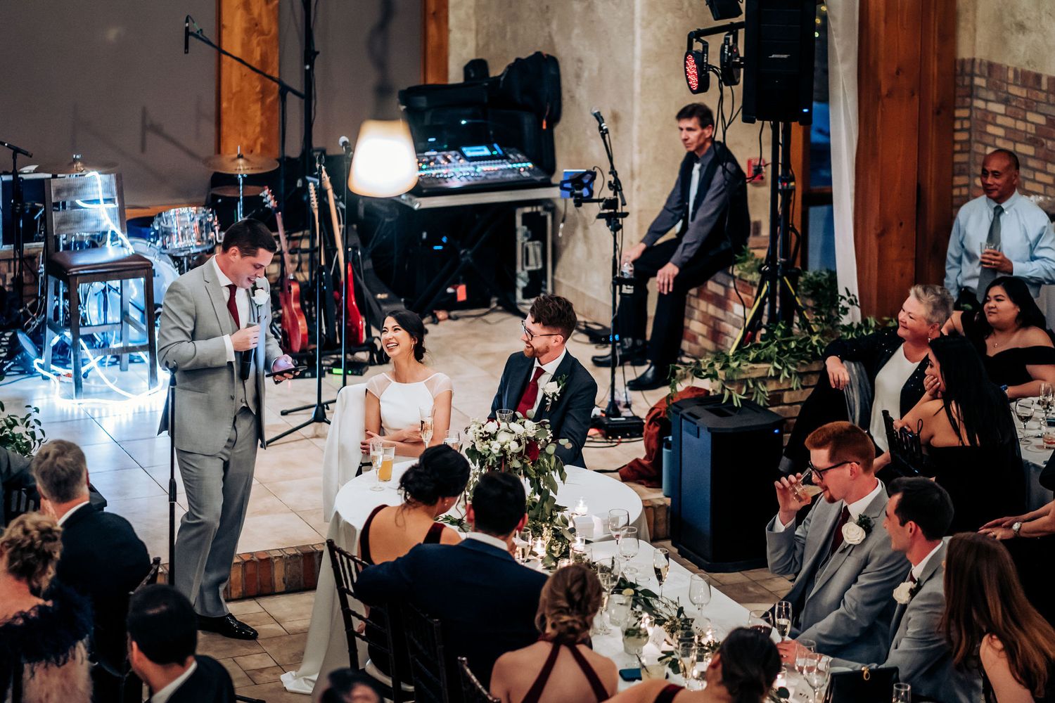 A speaker addresses wedding guests during a reception in an industrial-style venue with exposed brick walls and lighting.