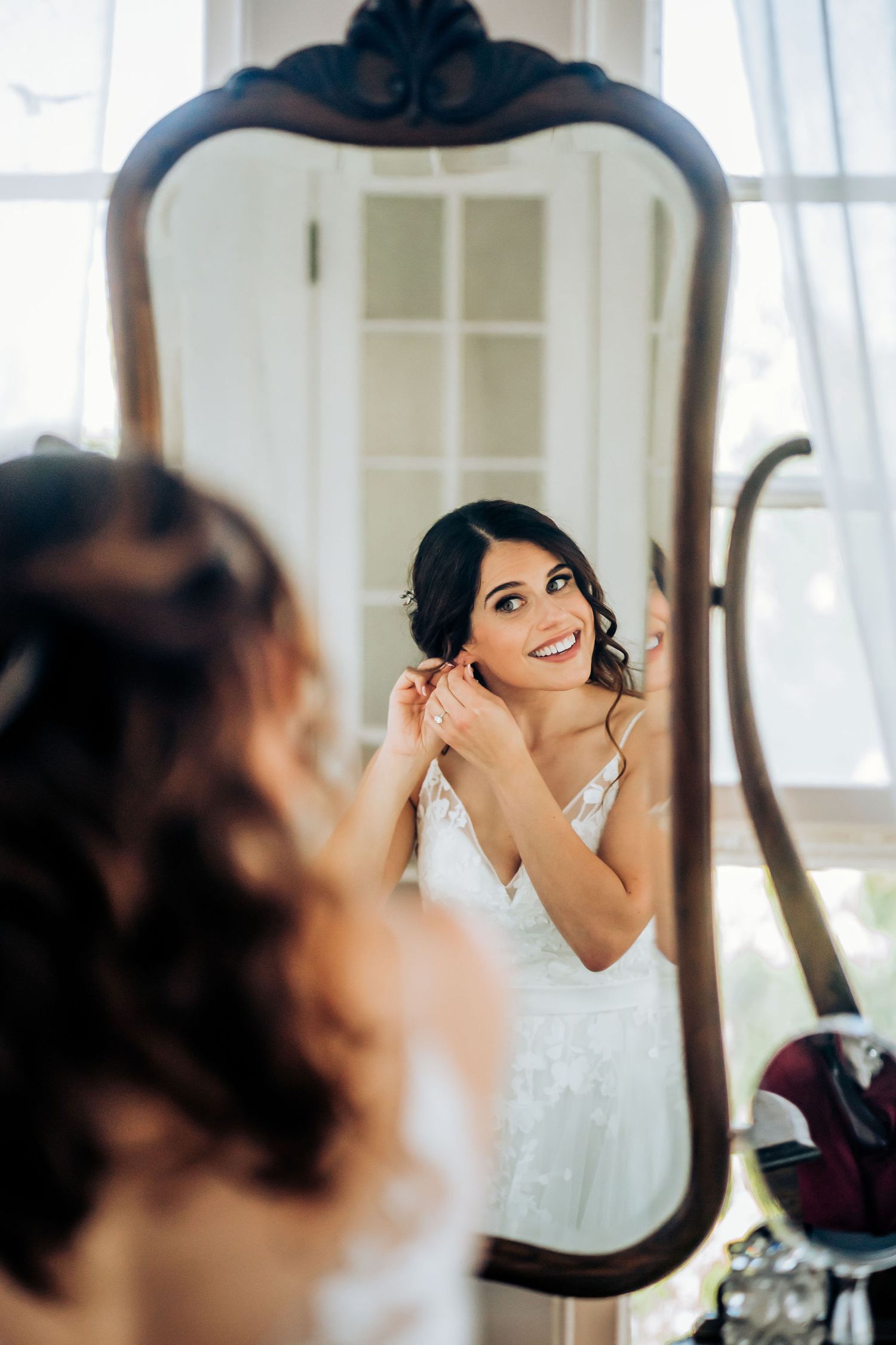 Reflection shows bride smiling while putting on jewelry in elegant mirror.