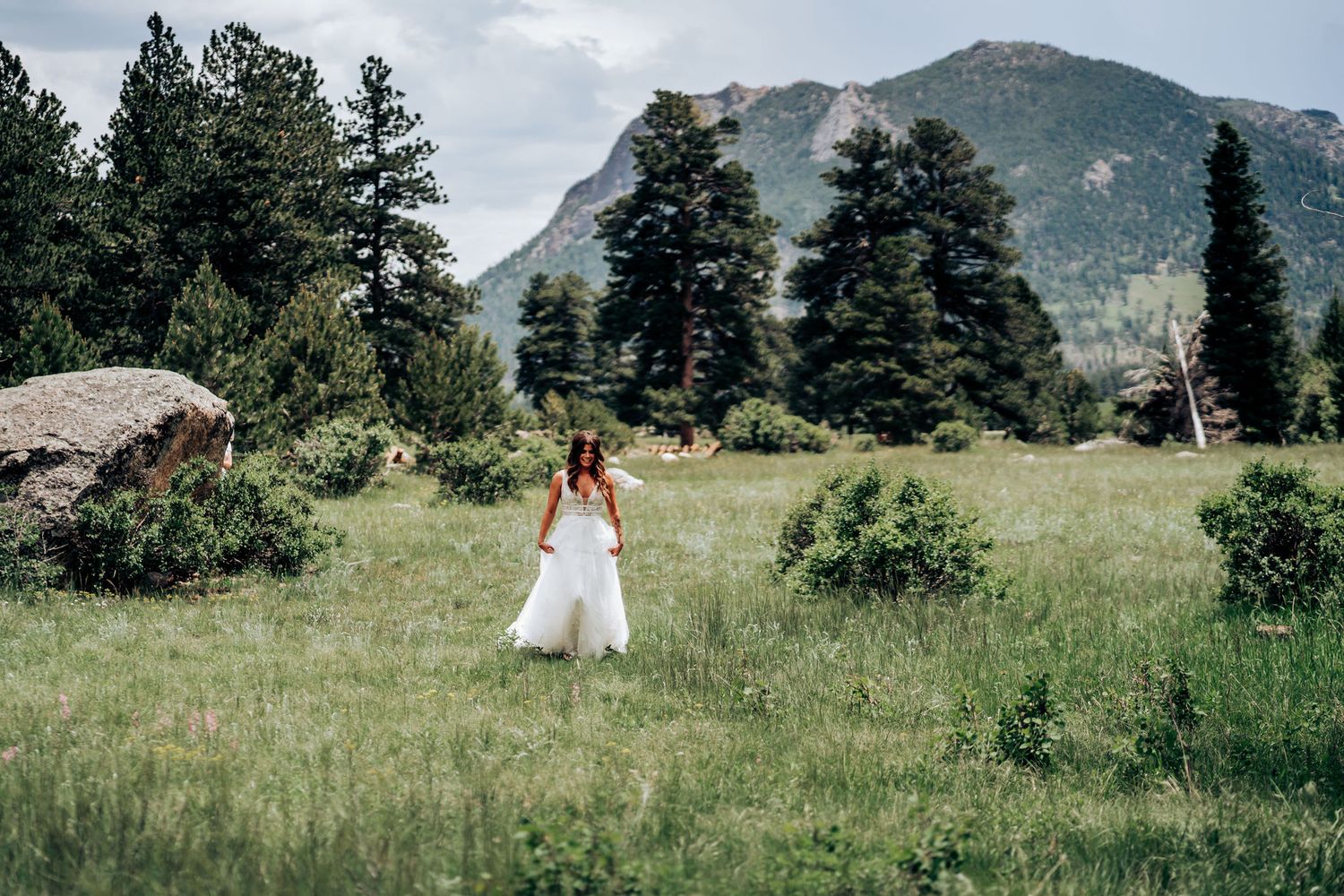 A bride in a flowing white dress walks through a mountain meadow with evergreen trees and peaks in background.