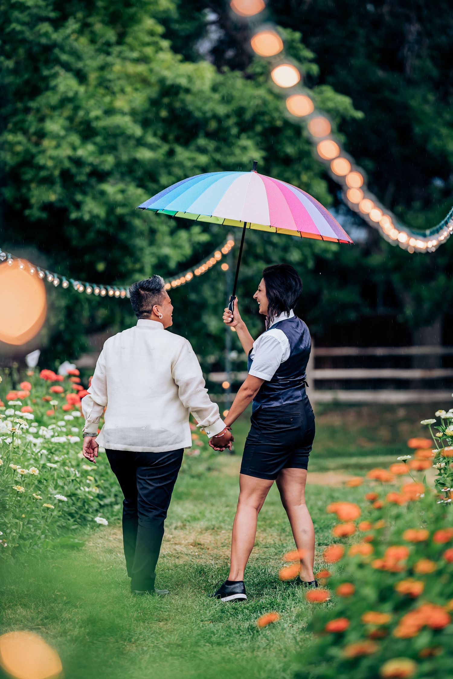 Two people stroll through a garden path under a colorful umbrella with string lights overhead.