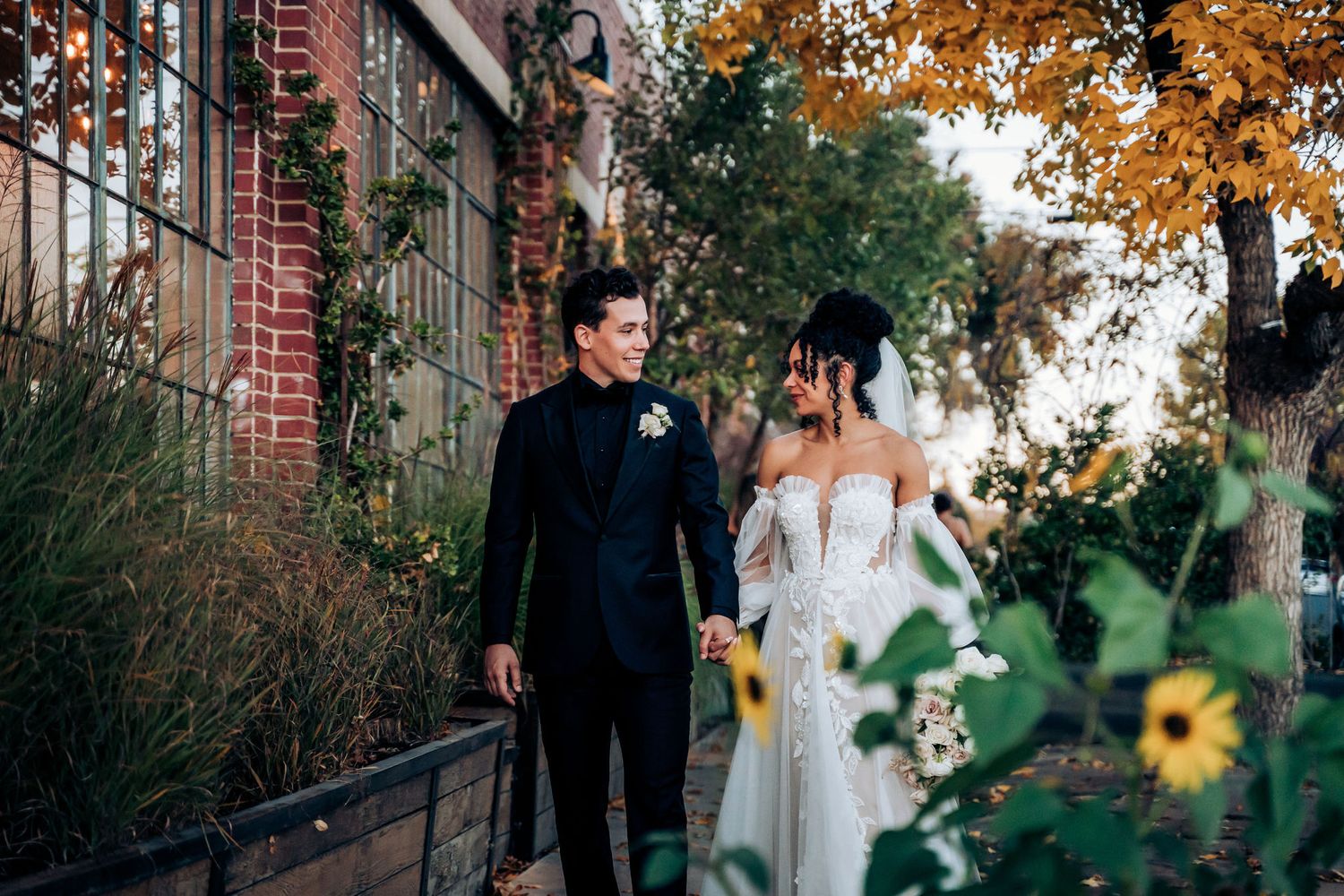 A wedding couple walks along a brick building path with autumn trees and flowers in the background.