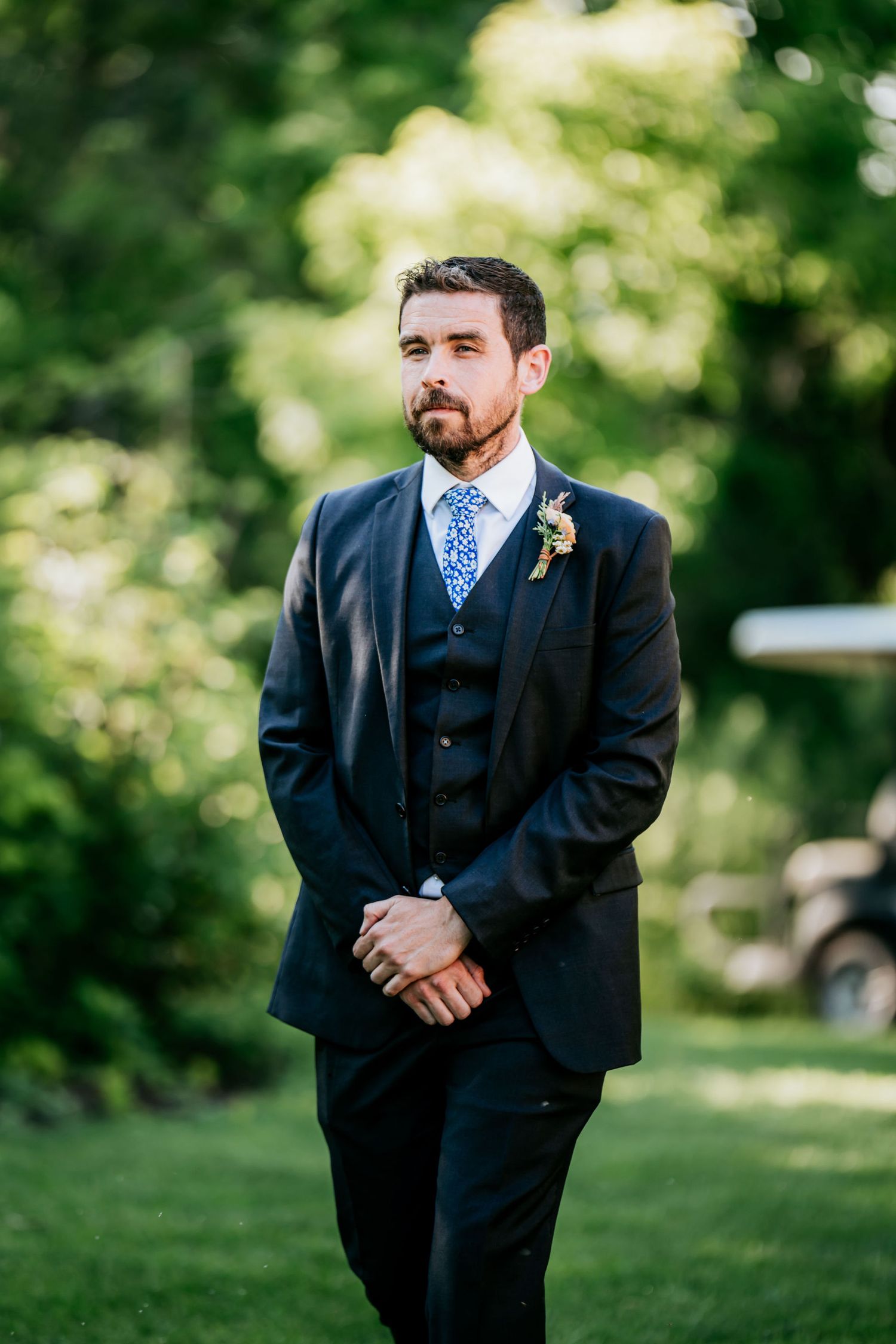 A formal portrait of someone in a dark three-piece suit with a blue tie outdoors.