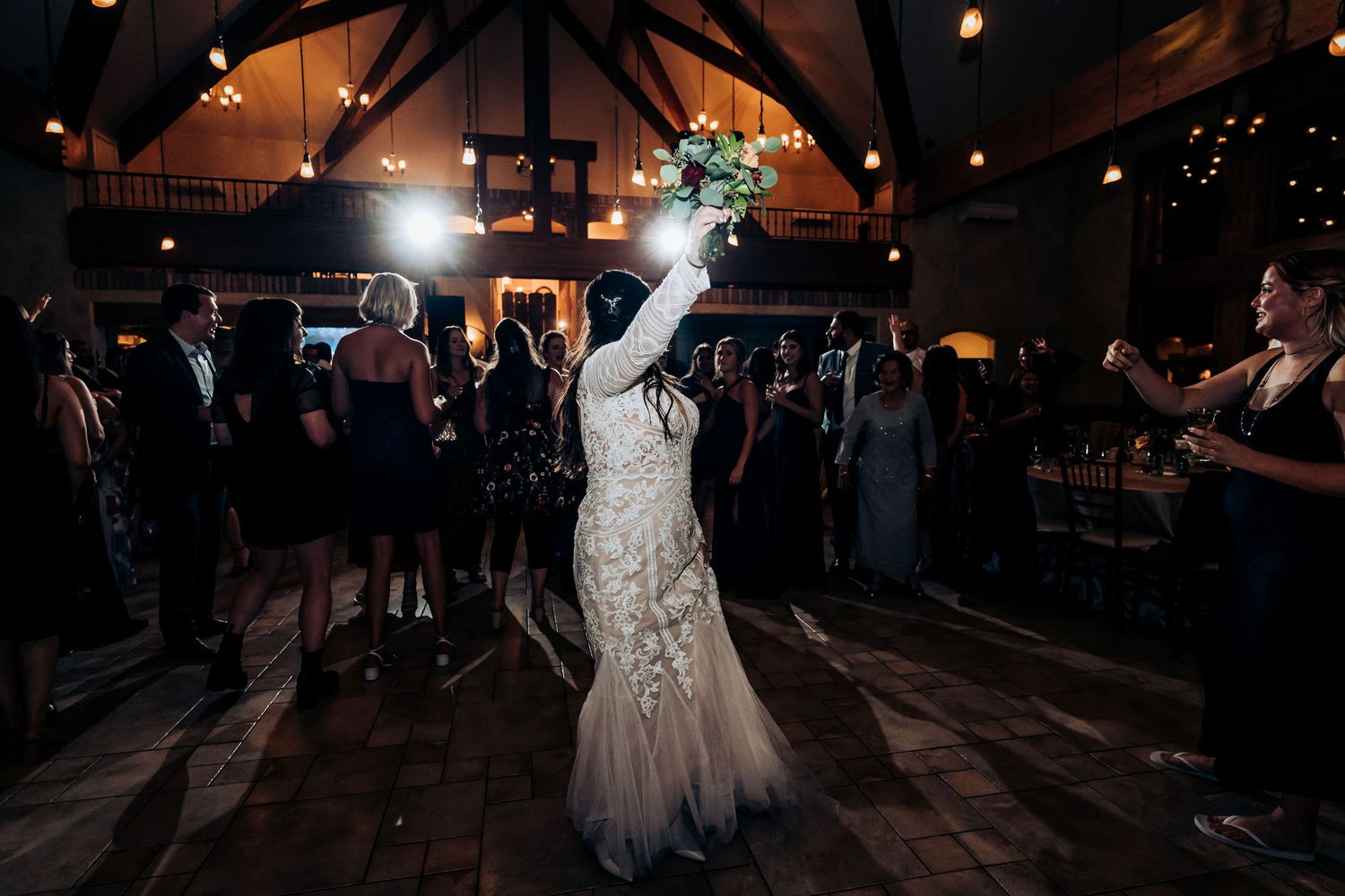 Person in lace gown raises bouquet while dancing under venue lighting.