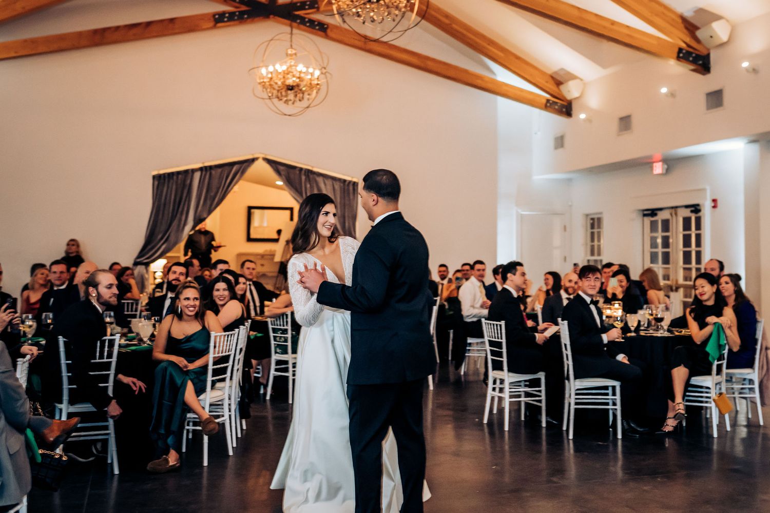 A couple shares their first dance in an elegant reception hall with exposed wooden beams and crystal chandeliers.
