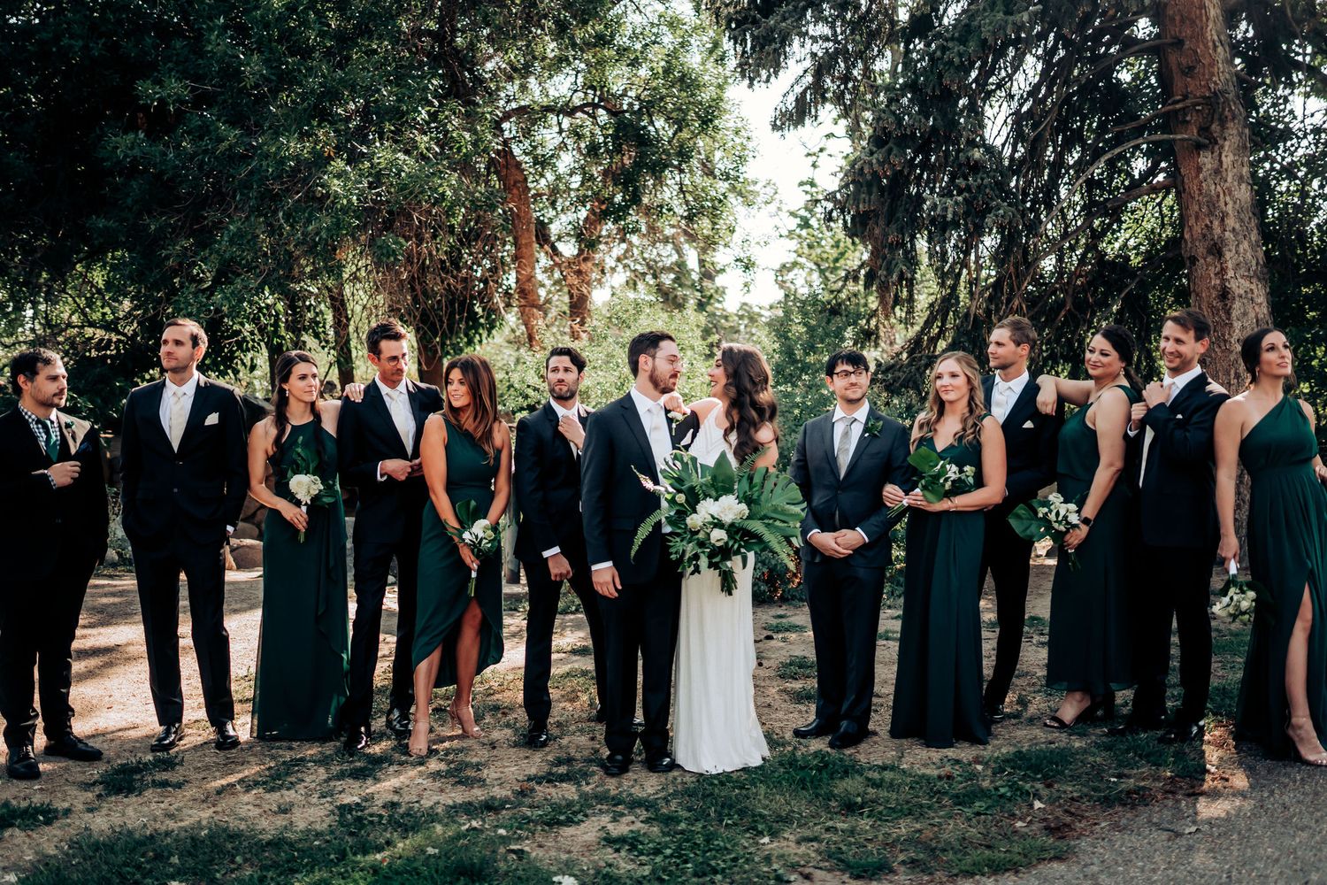 Wedding party lined up outdoors wearing coordinated black suits and dark green dresses.