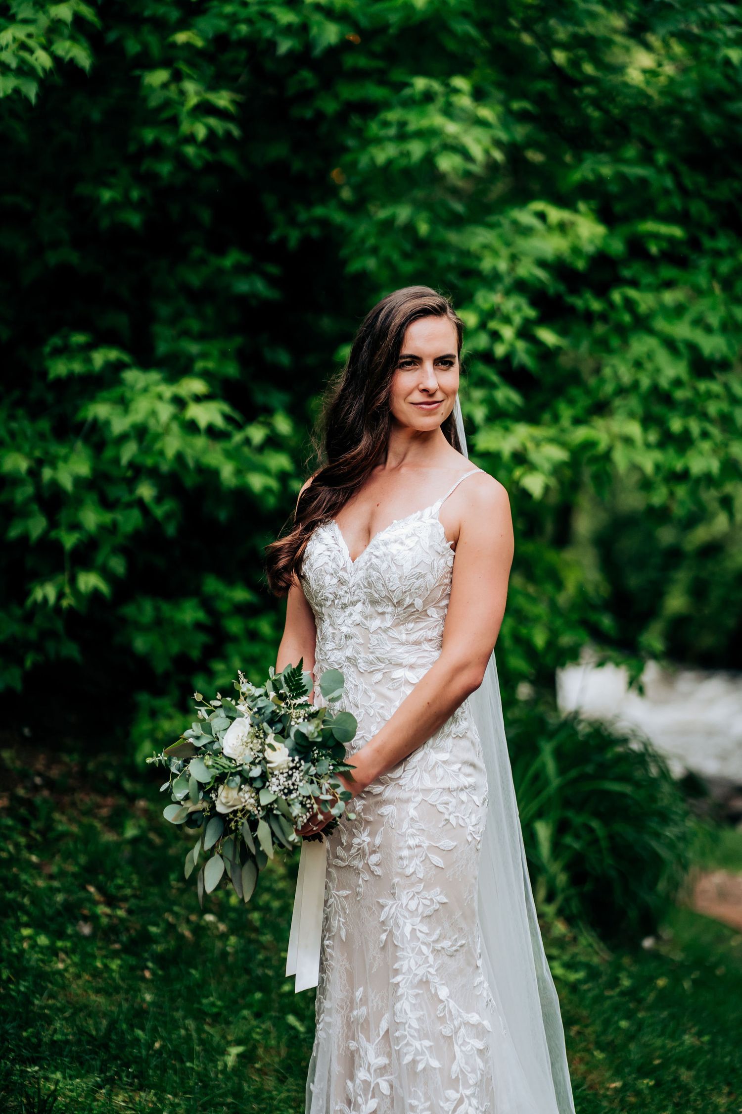 Bride in lace wedding gown holds greenery bouquet against natural garden backdrop.