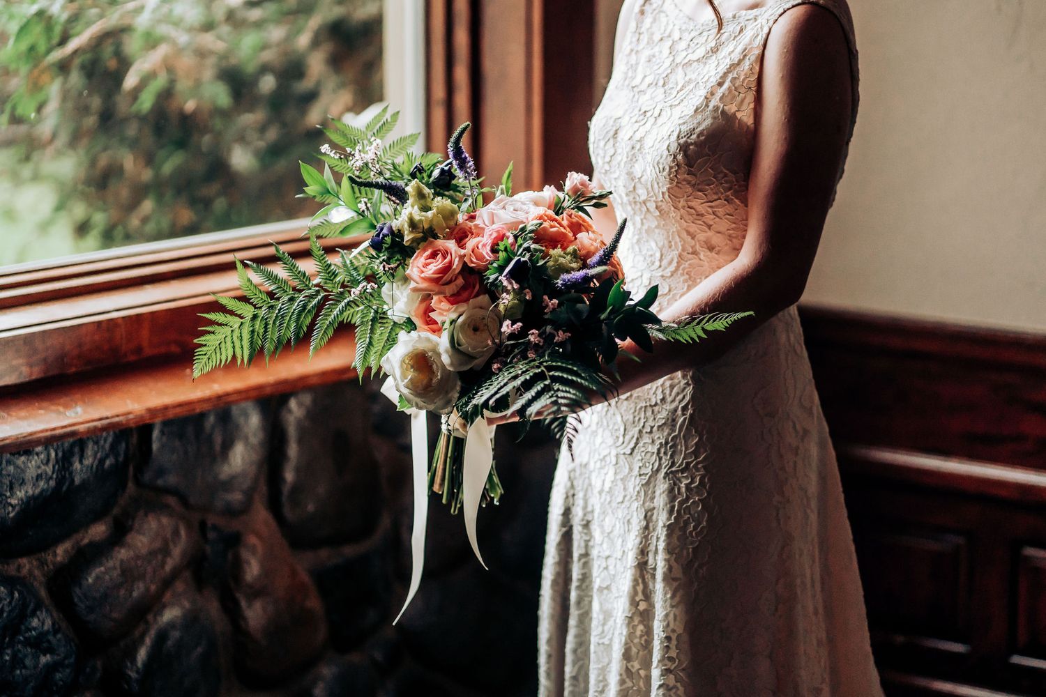 A delicate lace wedding dress and vibrant floral bouquet displayed near a stone wall and window.