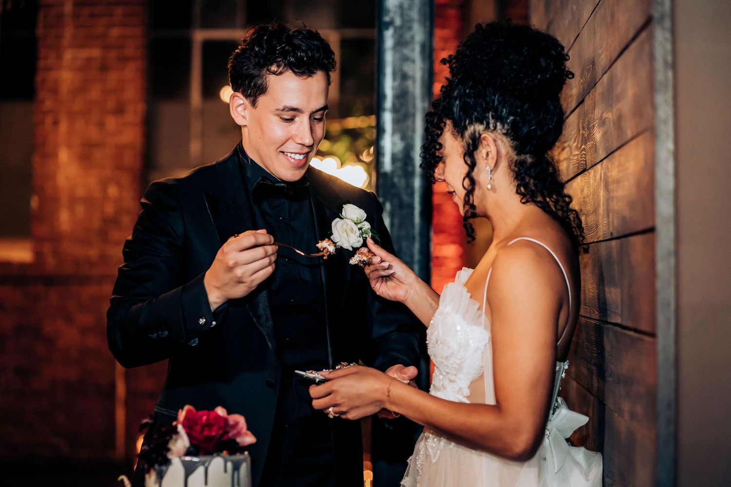 Newlyweds share a sweet cake tasting moment during their wedding reception against a brick wall backdrop.