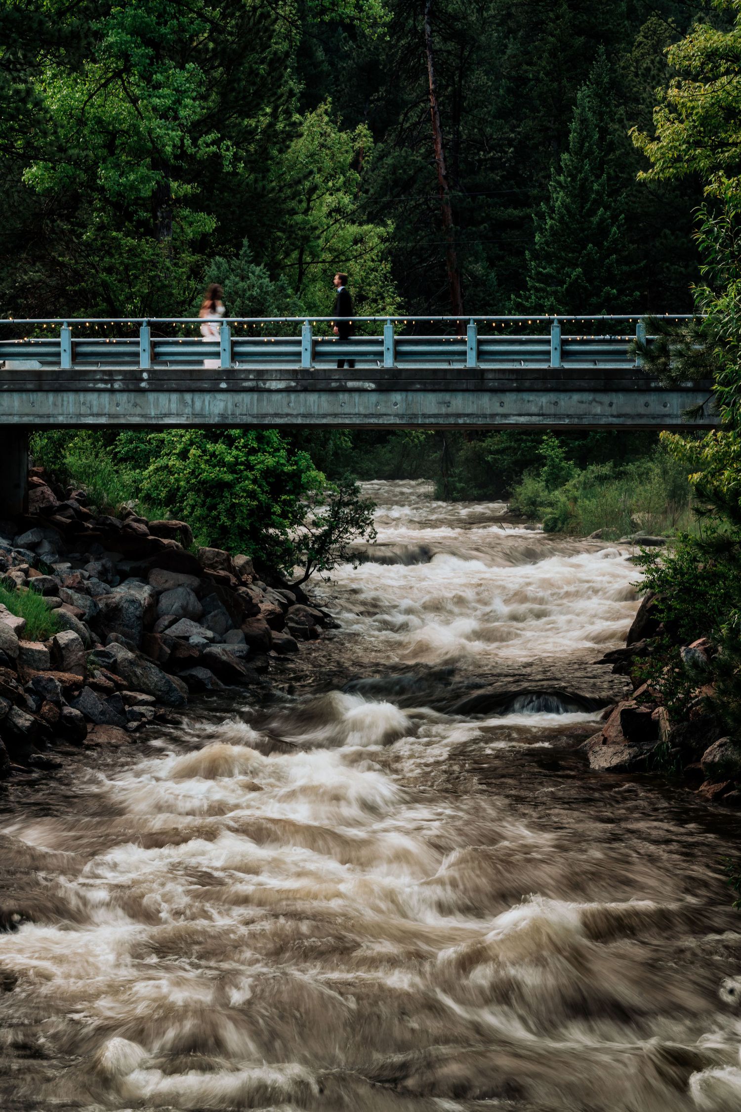 Small bridge spans over rushing whitewater rapids in a forested mountain setting.