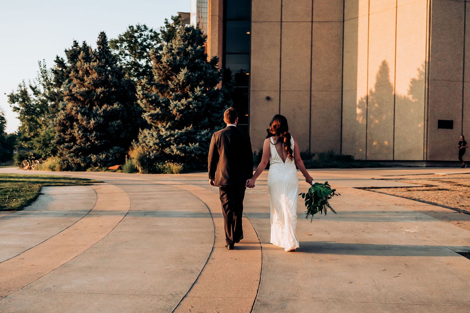 Newlywed couple walking together on a curved pathway holding a green and white bouquet.