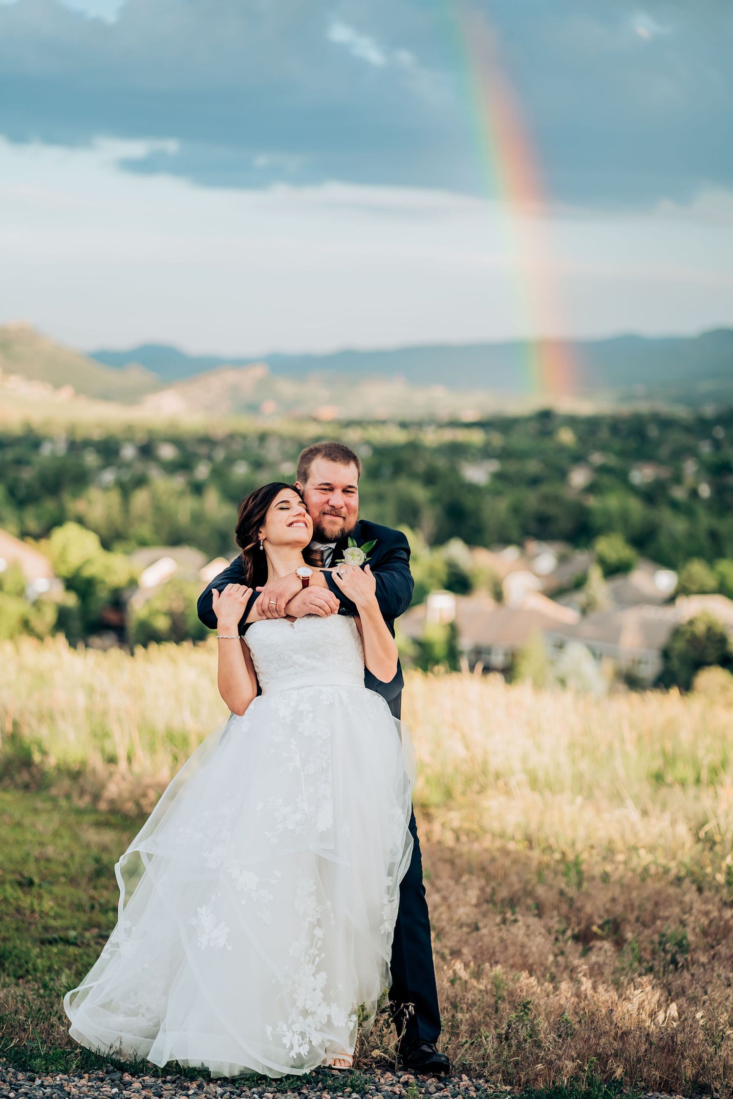 A couple shares a romantic moment in a meadow with a beautiful rainbow in the background on their wedding day.