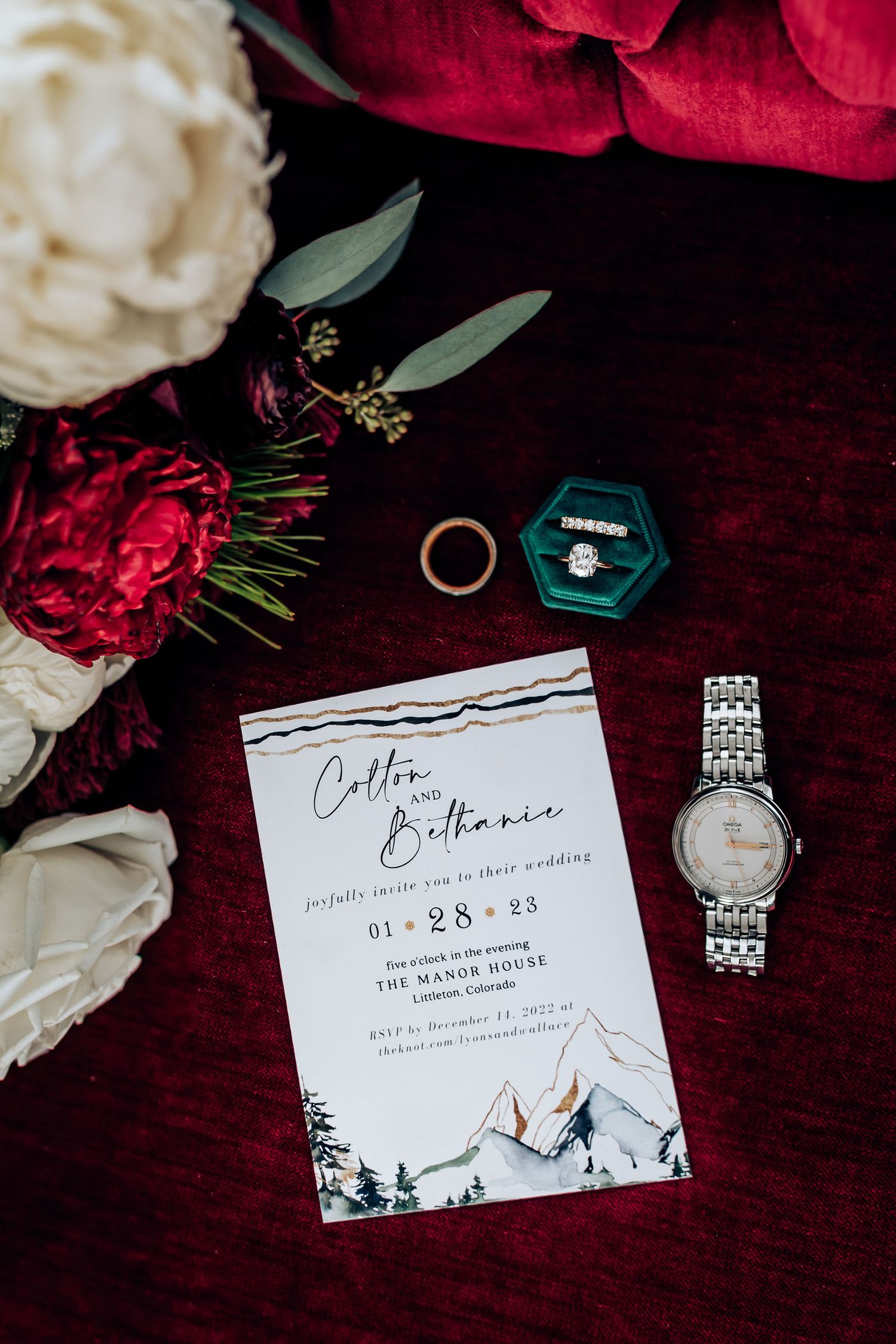 Wedding invitation with mountains design alongside rings, flowers, and watch on burgundy velvet.