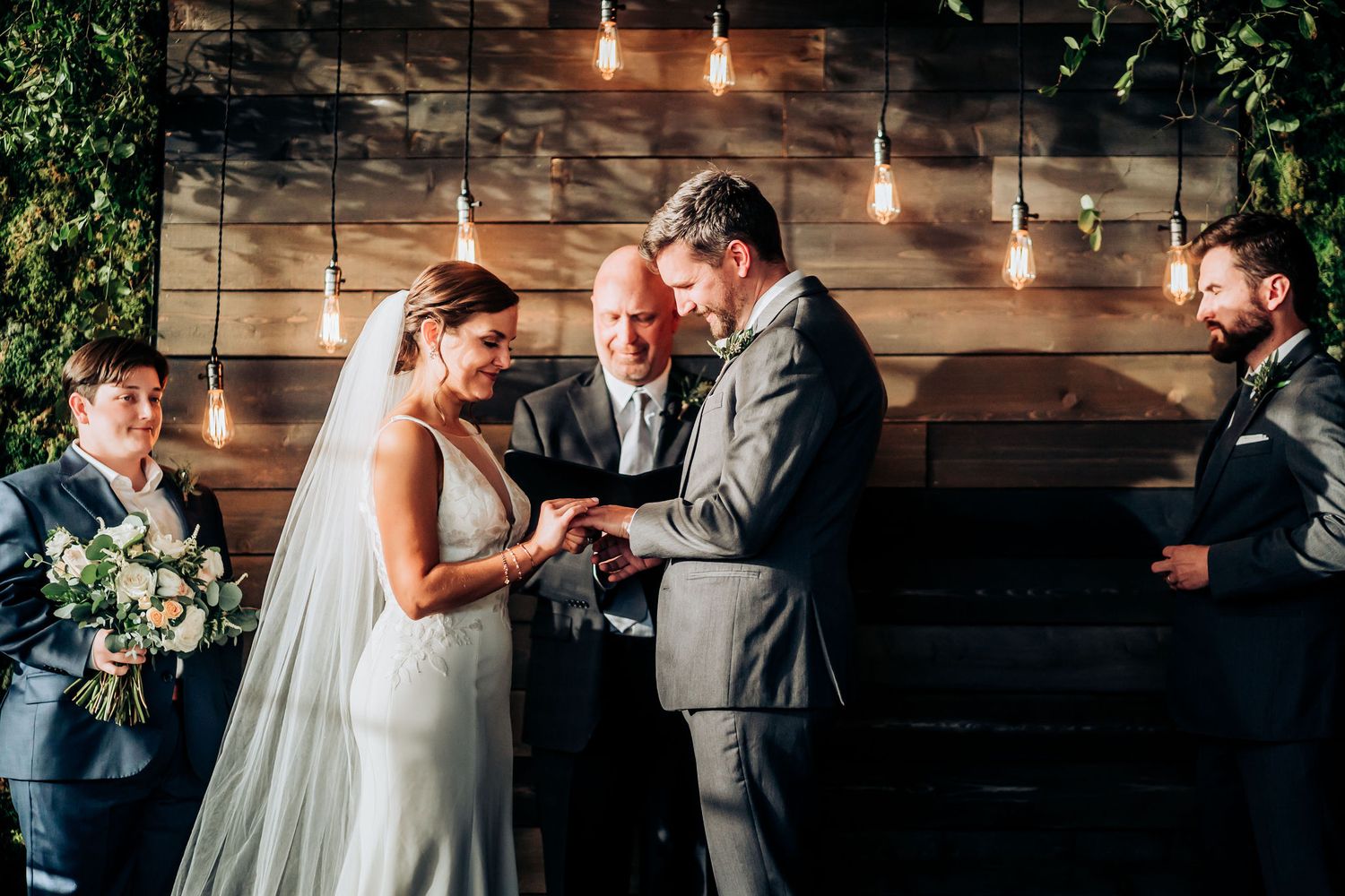 Wedding ceremony under hanging lights and greenery with rustic wood backdrop.