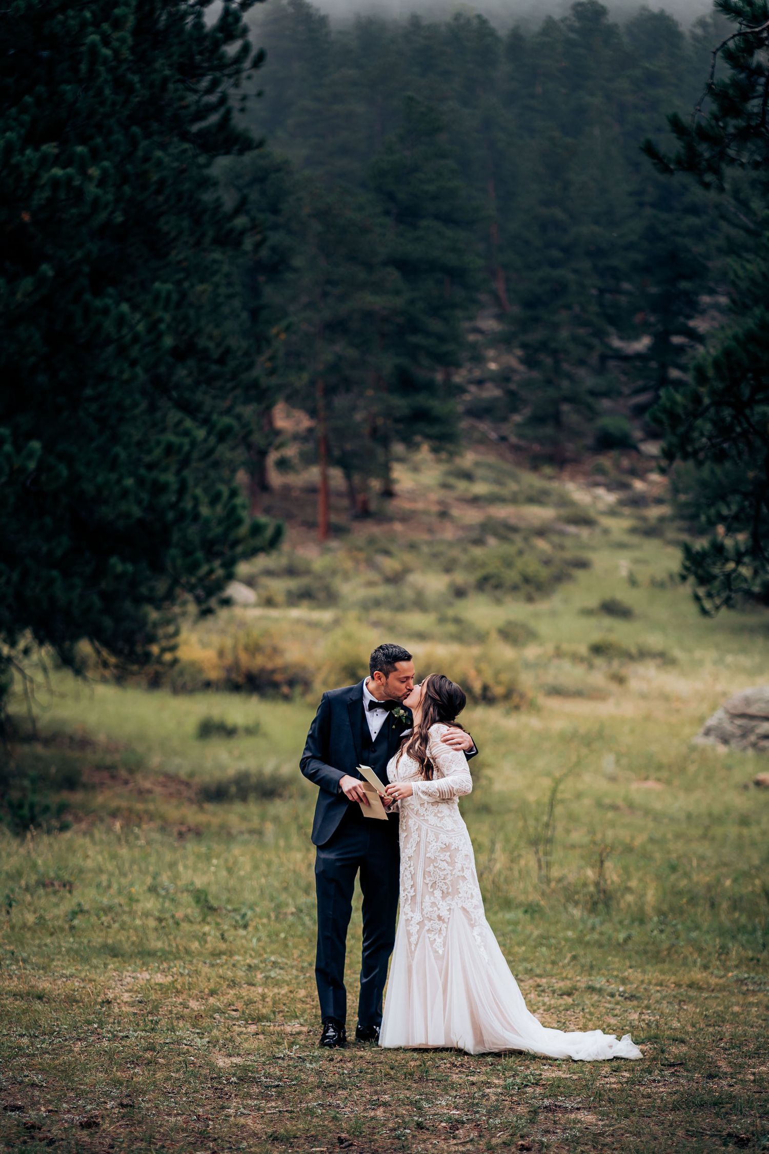 A couple shares an intimate moment in formal attire against a misty mountain backdrop.
