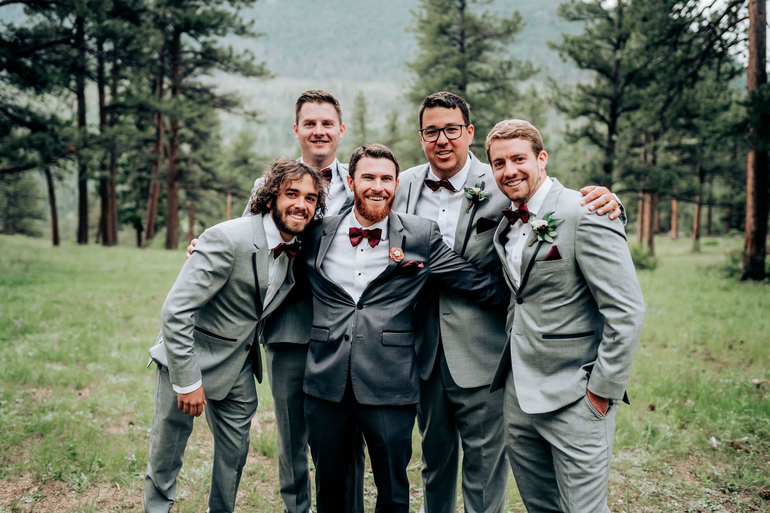 Groomsmen share a candid moment laughing together in gray suits against forest backdrop.