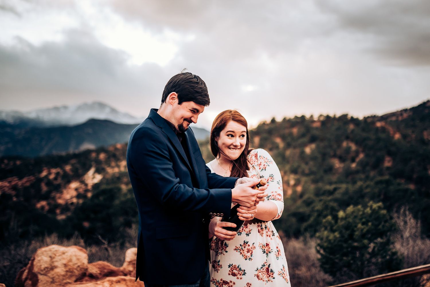 A couple in formal attire embraces on a rocky overlook with mountain views.
