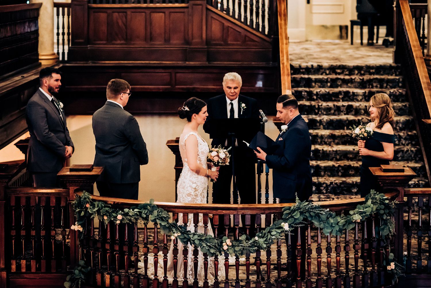 Wedding ceremony being held in a historic venue with wooden staircases and ornate banisters.