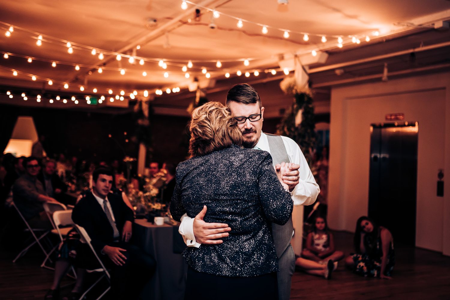 Two figures embrace during a dance at an indoor celebration with string lights overhead.