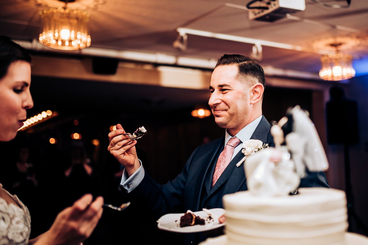 A couple shares wedding cake during their reception under elegant chandelier lighting.