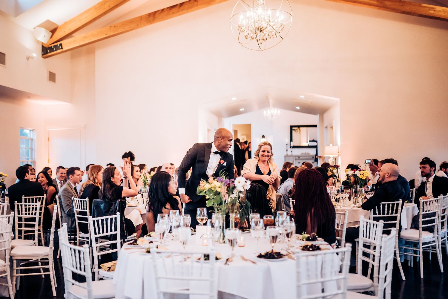 Wedding reception with white table settings and elegant chandelier in bright indoor venue.