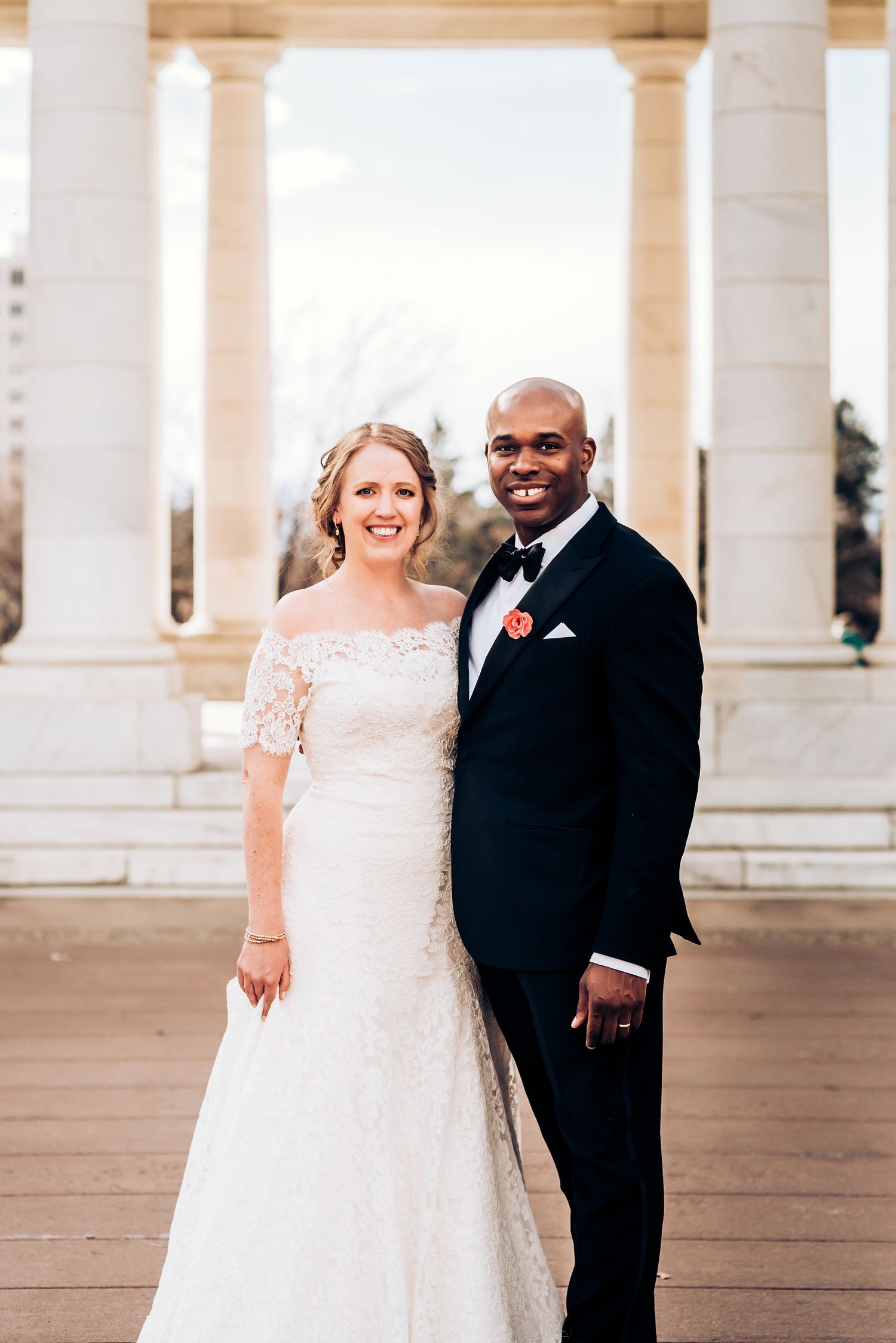 Wedding couple poses elegantly in front of white stone columns at sunset.