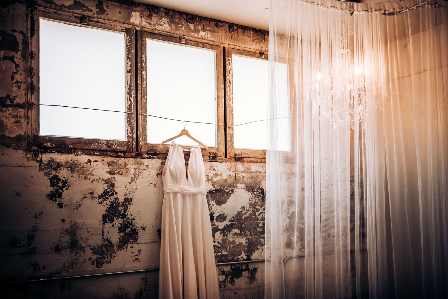 A simple white wedding dress hangs in front of sunlit windows in a rustic room.