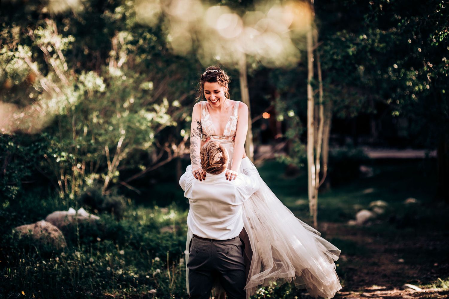 A romantic moment in a sunlit forest as a couple shares a playful lift wearing formal wedding attire.
