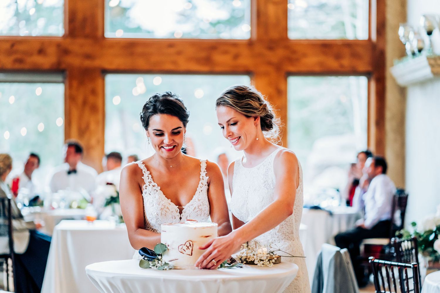 Two people in white dresses sharing a joyful moment while cutting cake at an elegant wedding reception.