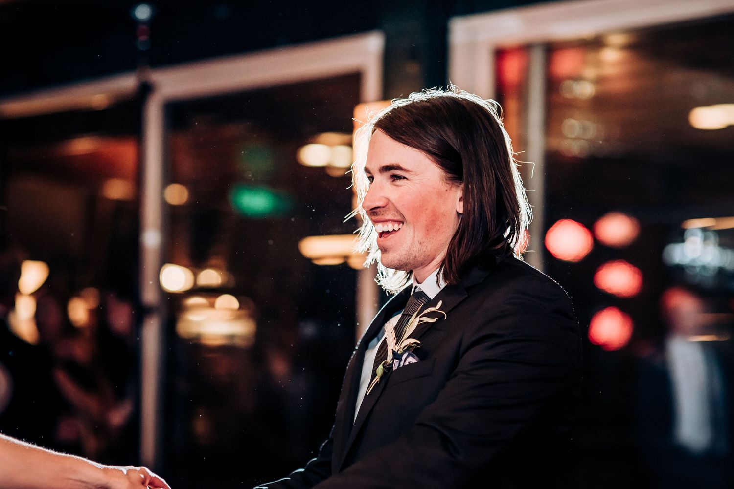A person in formal attire laughs joyfully during a wedding celebration with bokeh lights in background.