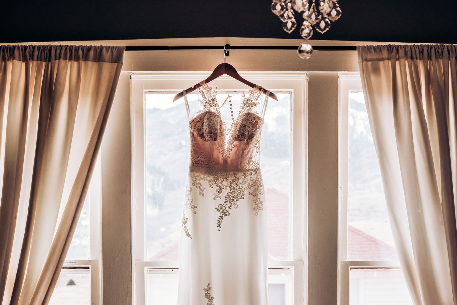 A delicate lace wedding gown suspended in front of a window with flowing cream-colored curtains.