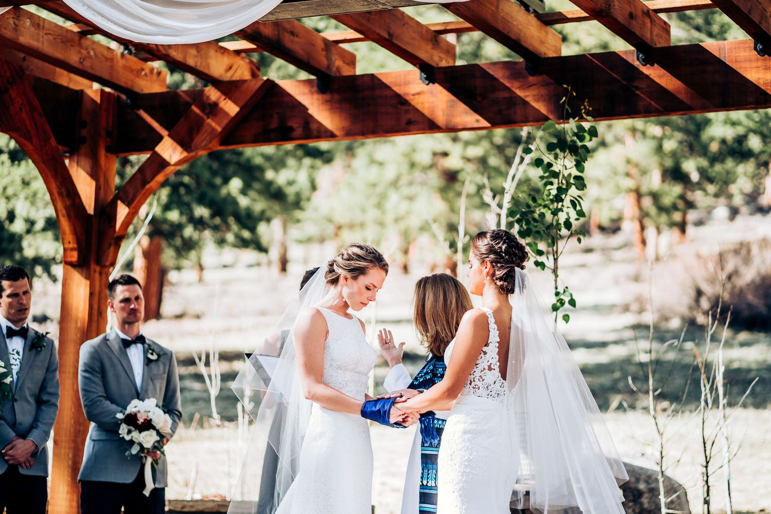 A wedding ceremony under a wooden pergola with an officiant in blue stole conducting the service.