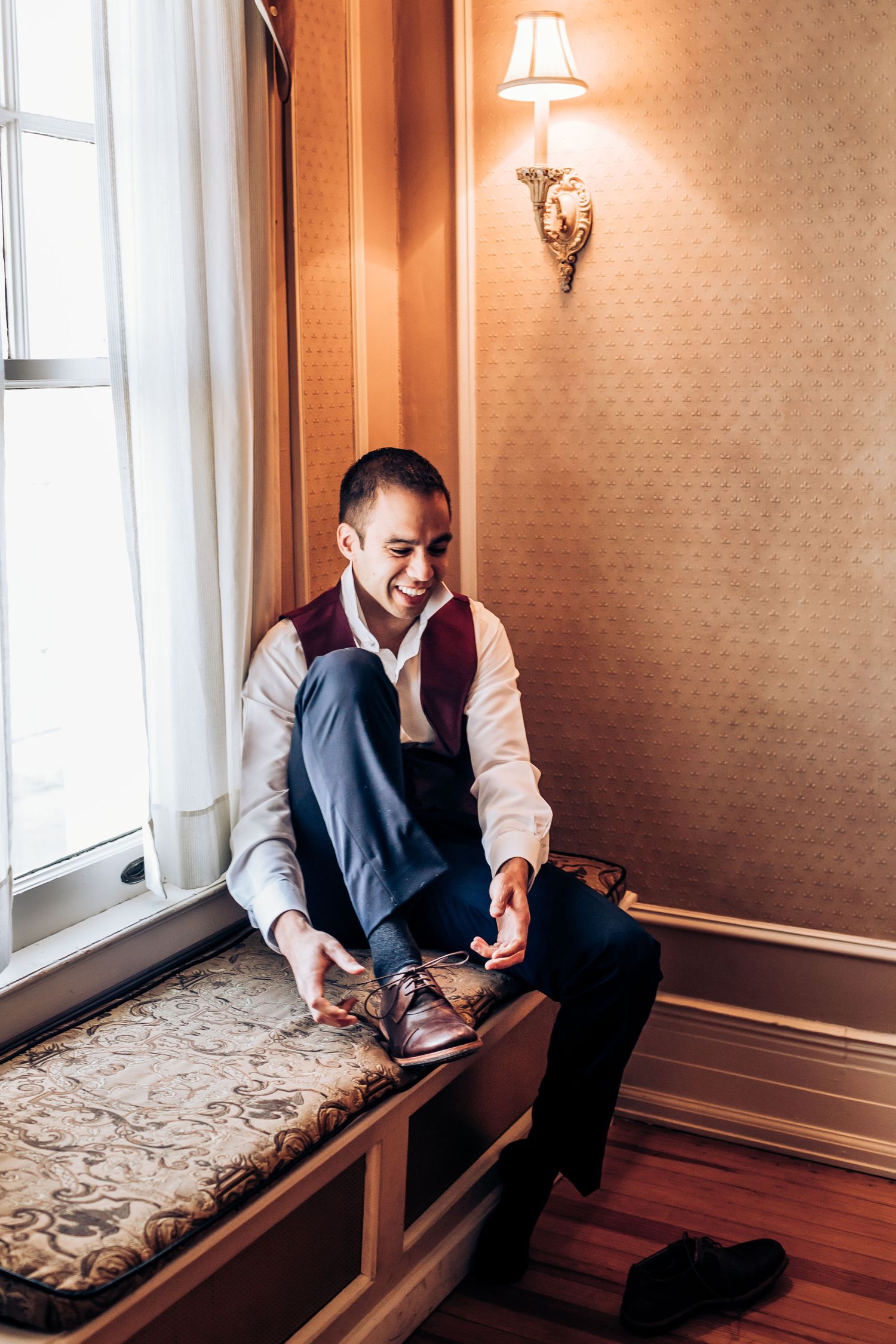 Man in formal attire puts on dress shoes while seated by window.